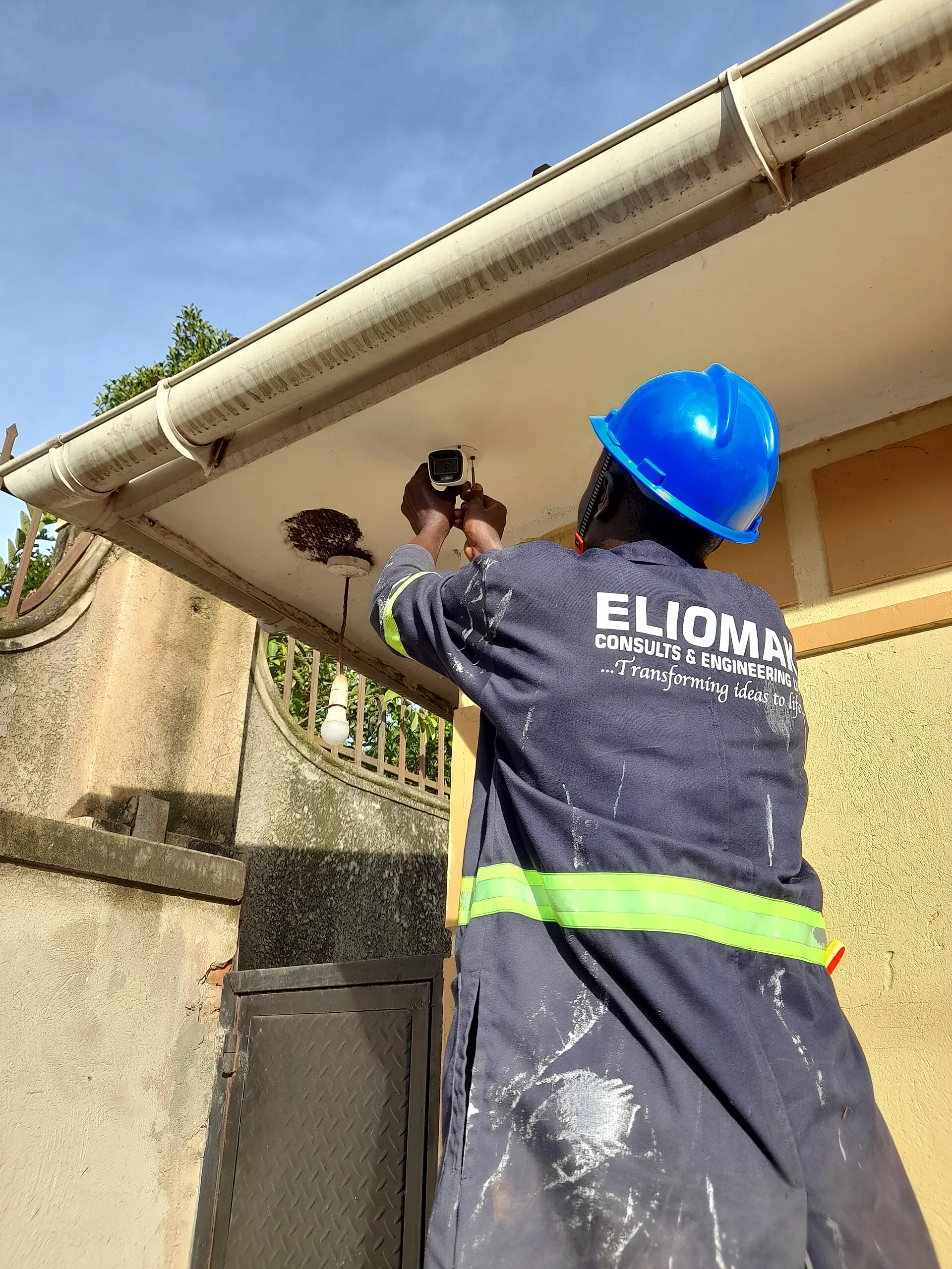 Technician in a blue hard hat and reflective overalls screwing a white bullet camera into the underside of a roof eave