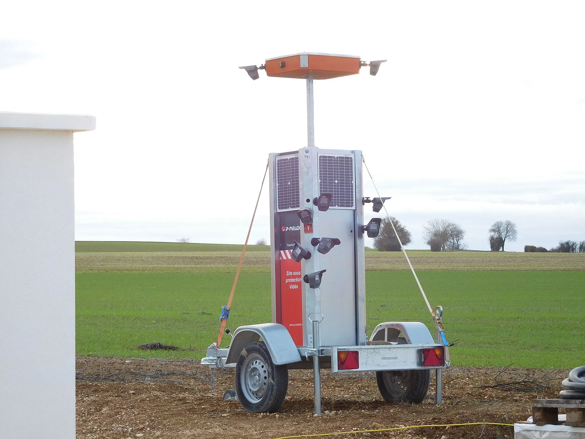 Trailer-mounted tower with two solar panels and several small cameras on arms standing in a farm field