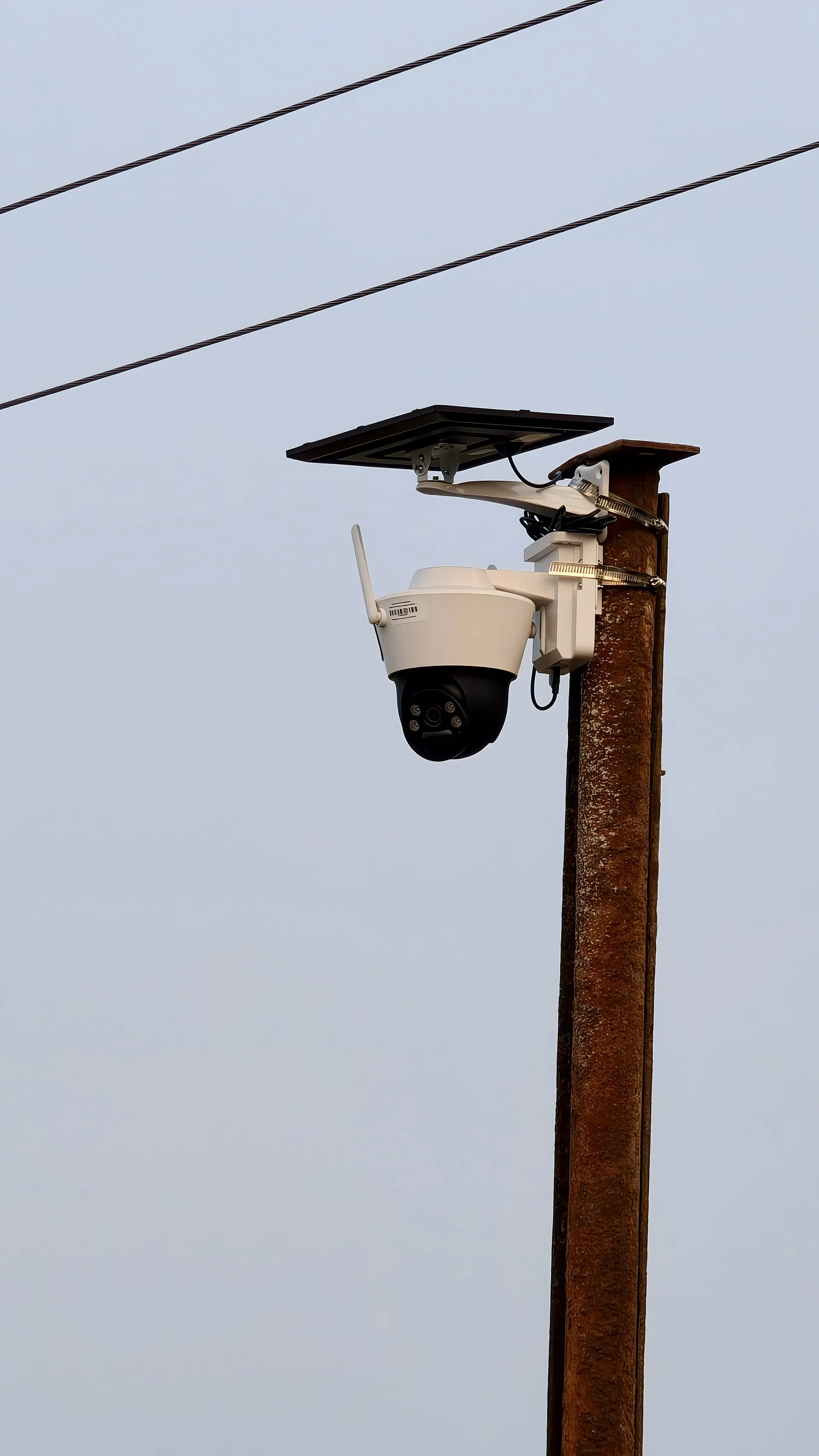 White pan and tilt camera with a solar panel above it, clamped to a rusted pole with wires behind it