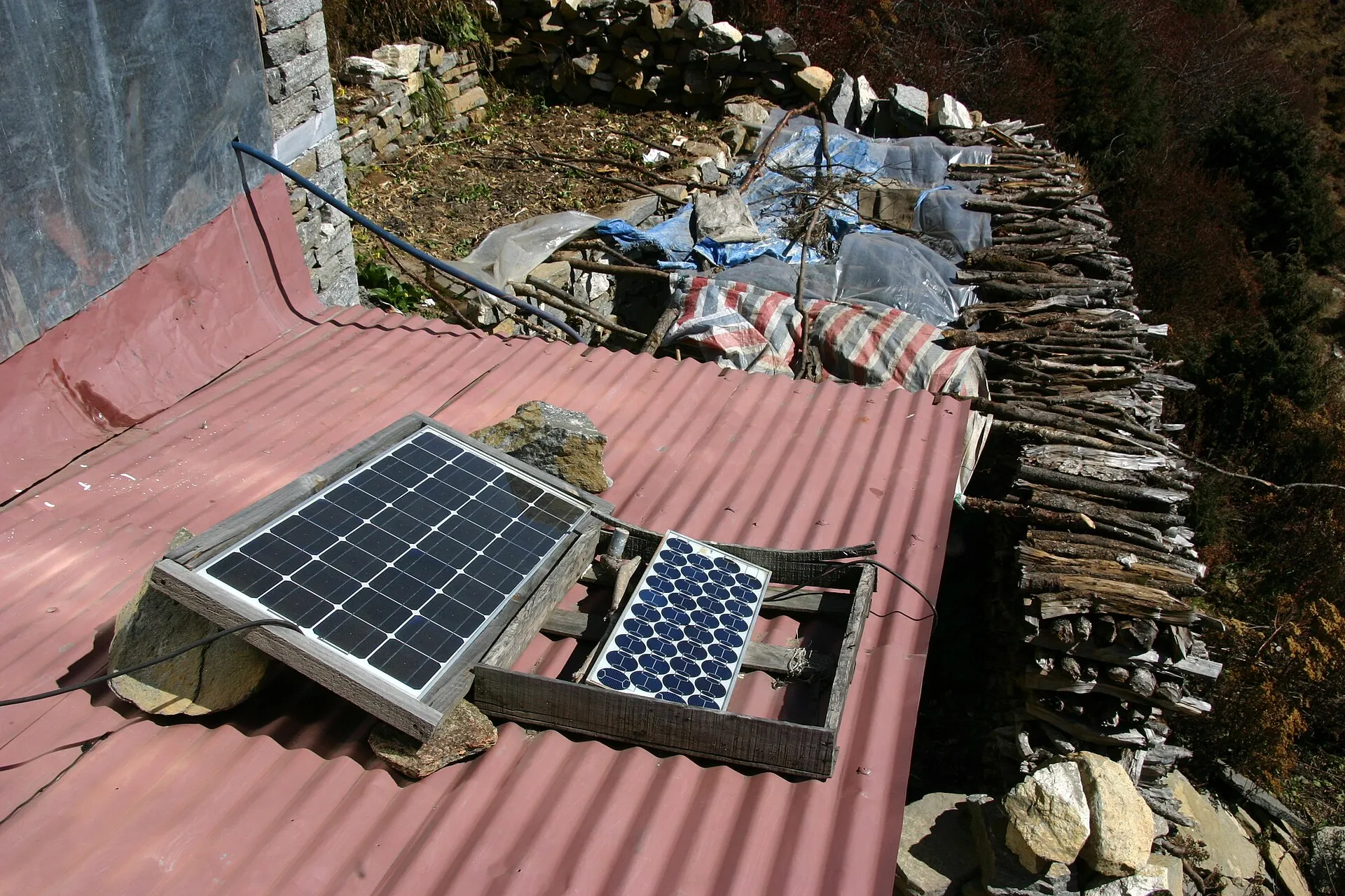 Two small solar panels in wooden frames resting on a red corrugated metal roof beside stacked firewood on a hillside